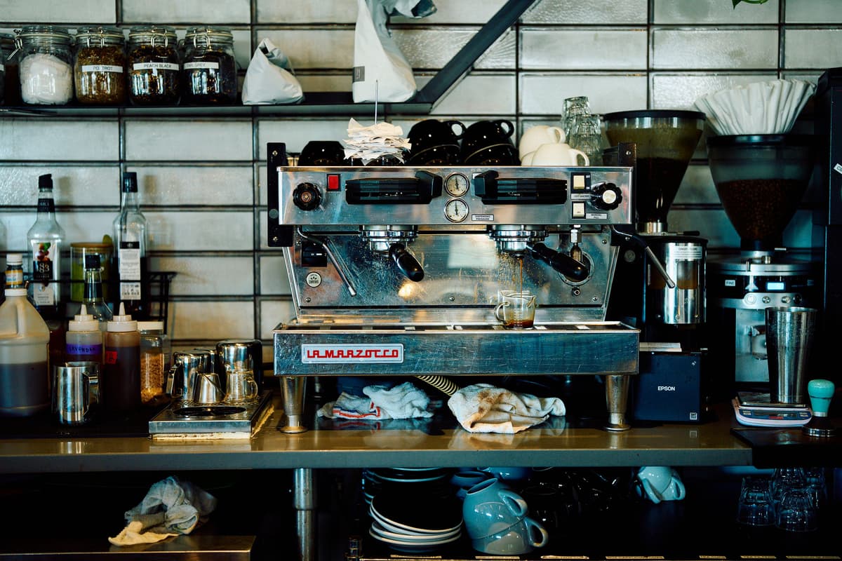 A busy café counter with a La Marzocco espresso machine, coffee cups, jars, and syrups. The scene conveys a bustling, well-used atmosphere.