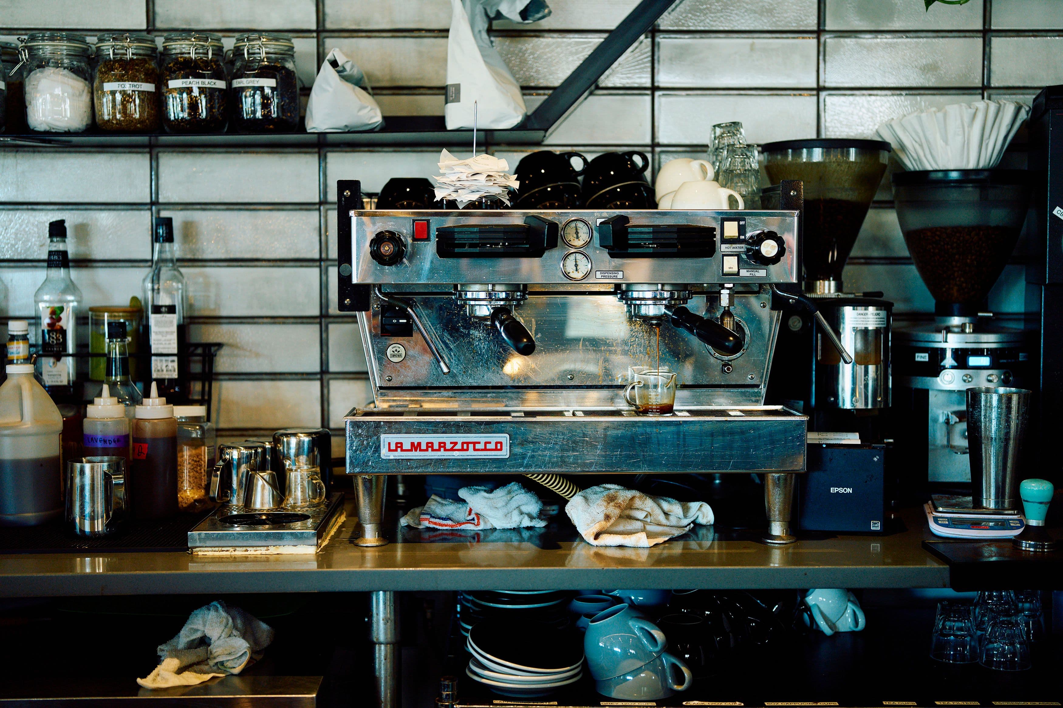 A busy café counter with a La Marzocco espresso machine, coffee cups, jars, and syrups. The scene conveys a bustling, well-used atmosphere.