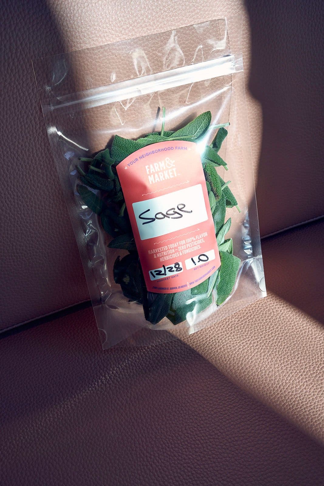 Resealable bag of fresh sage with a pink label reading “Farm & Market” and “Sage,” placed on light brown leather, sunlight casting a shadow.
