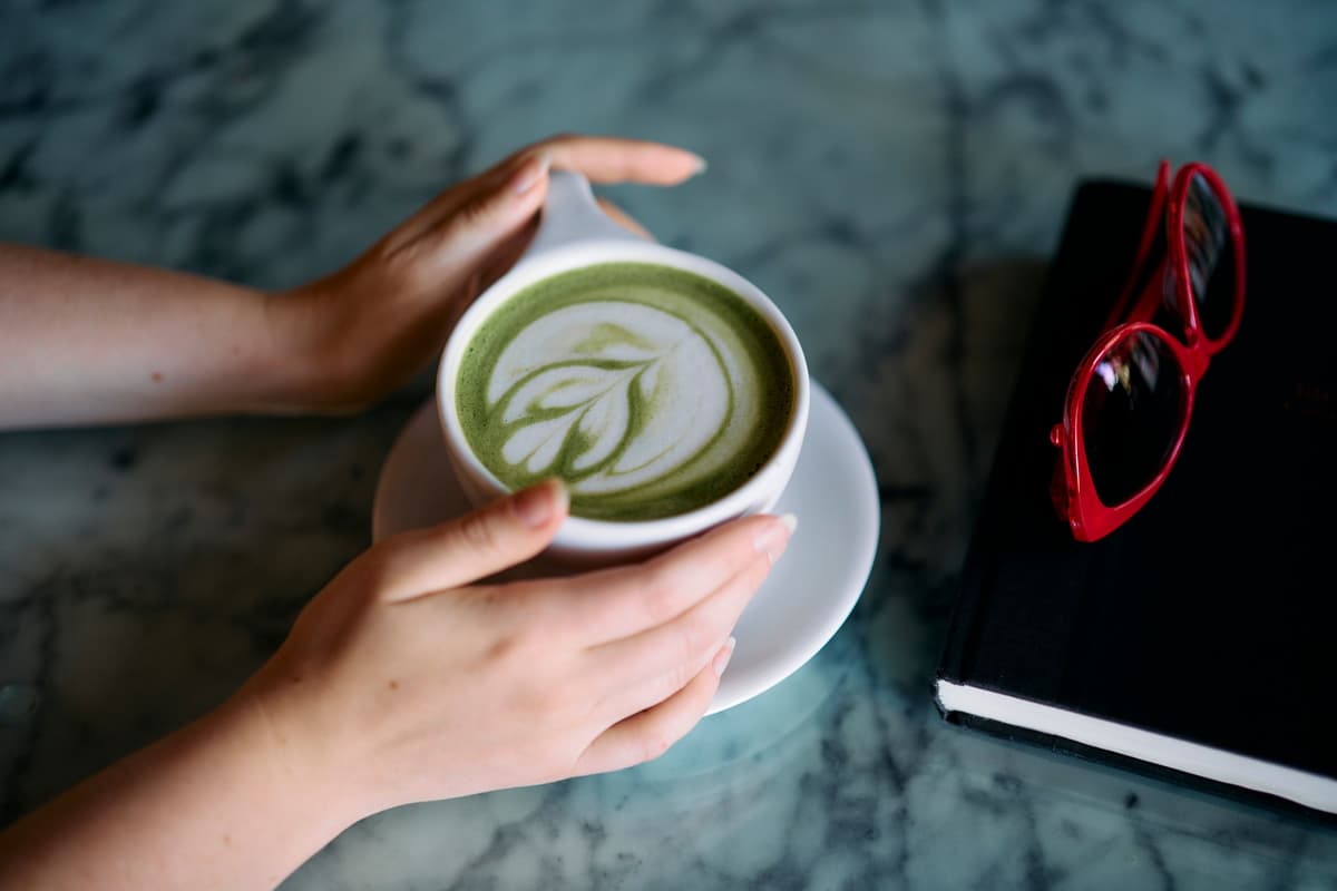 Two hands cradle a mug of matcha latte with leaf latte art on a marble table. A book with red sunglasses rests nearby, creating a cozy vibe.
