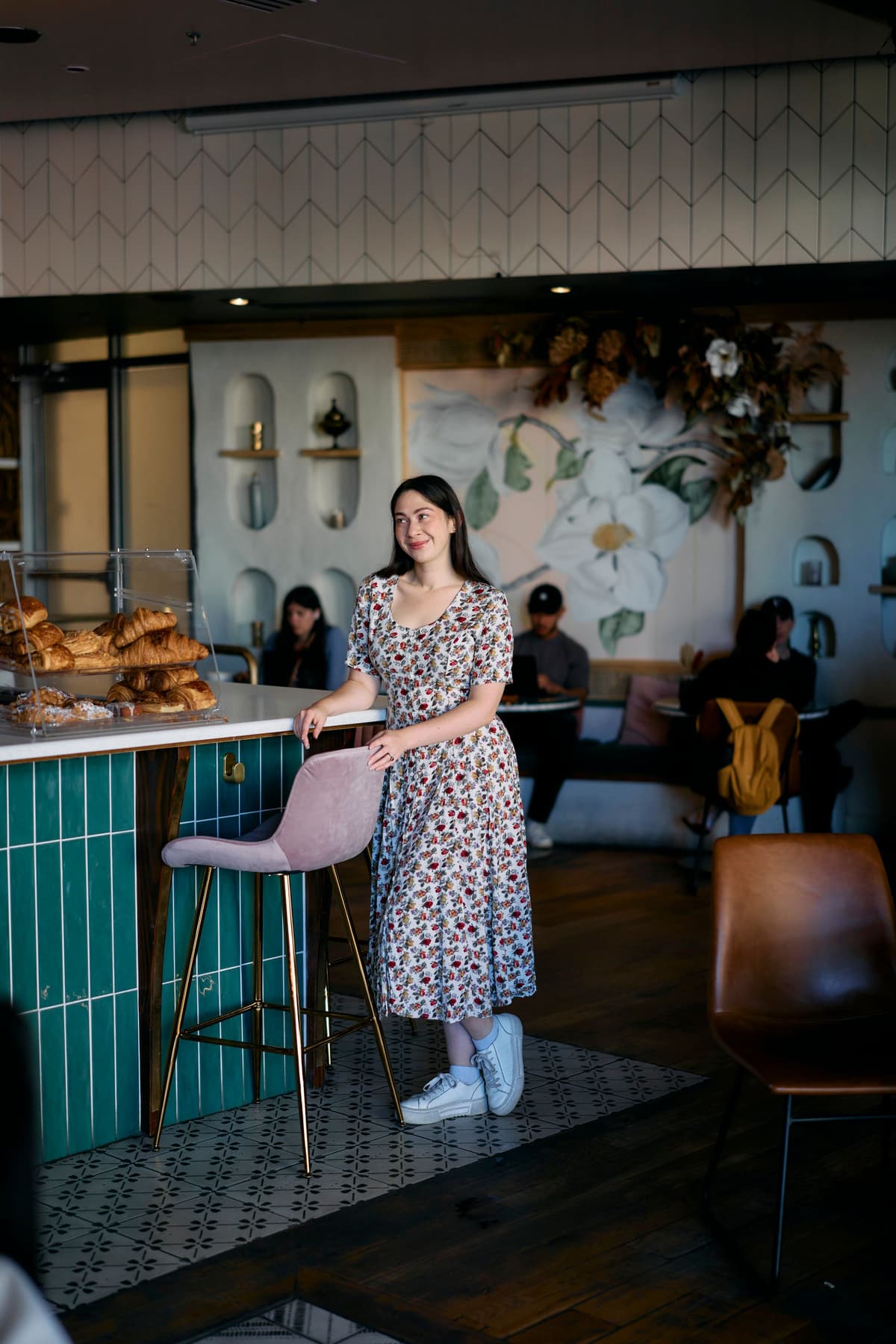 A woman in a floral dress stands cheerfully by a café counter with croissants. The background features a mural of flowers, teal tiles, and relaxed patrons.