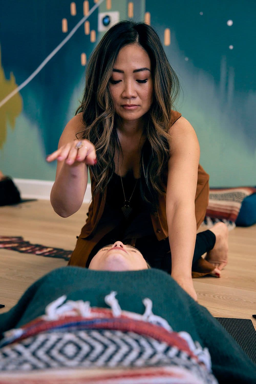A woman performs energy healing over another person lying on a mat, set in a calm room with a blue and green mural. The atmosphere is peaceful and focused.