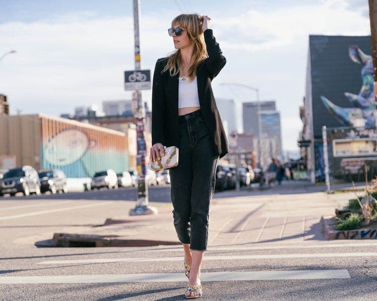 Woman in a black blazer and jeans walking confidently on a city street. She holds a patterned clutch, under a blue sky, conveying a chic urban vibe.