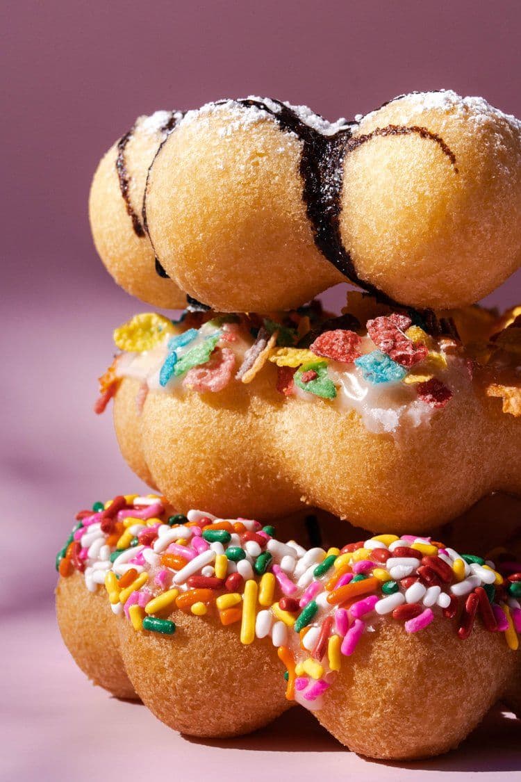 Close-up of a stack of mochi doughnuts with vibrant toppings. The top is drizzled with chocolate, the middle has colorful cereal bits, and the bottom is covered with rainbow sprinkles on a pink background.