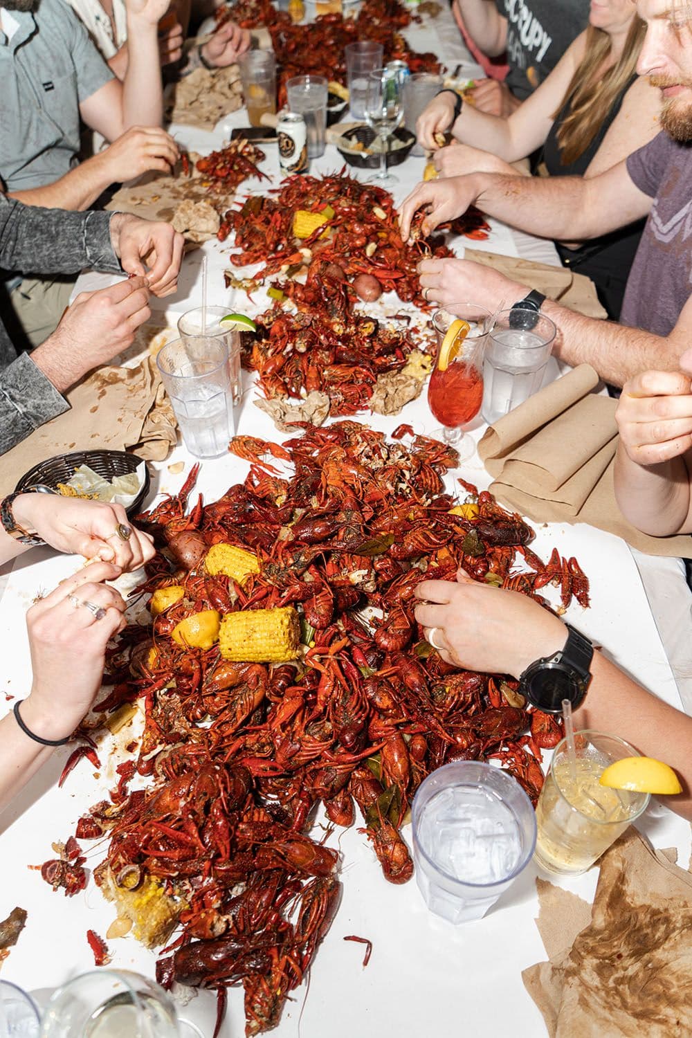 People gathered around a table covered with cooked crawfish, corn, potatoes, drinks, and napkins, eating and socializing at a crawfish boil.